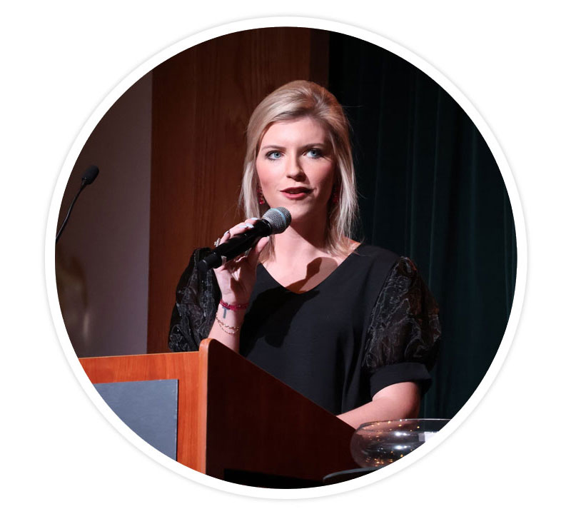 A woman with blonde hair holding a microphone and standing behind a podium talking, wearing a black shirt.